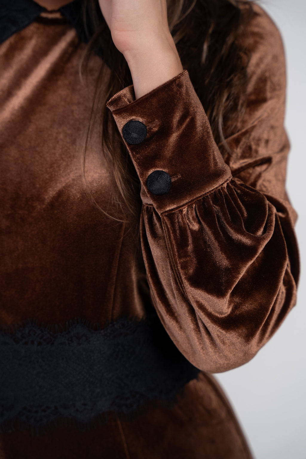 Close-up of a brown velvet long-sleeve top or dress with black buttons on the cuff, gathered sleeves, and a black lace band near the waist, shown on a person against a light gray background.