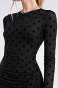 A close-up of a model wearing a black long-sleeve dress with a sheer mesh fabric and a black polka dot pattern. The dress features a gathered ruched waist and a sparkling rhinestone-trimmed neckline.
