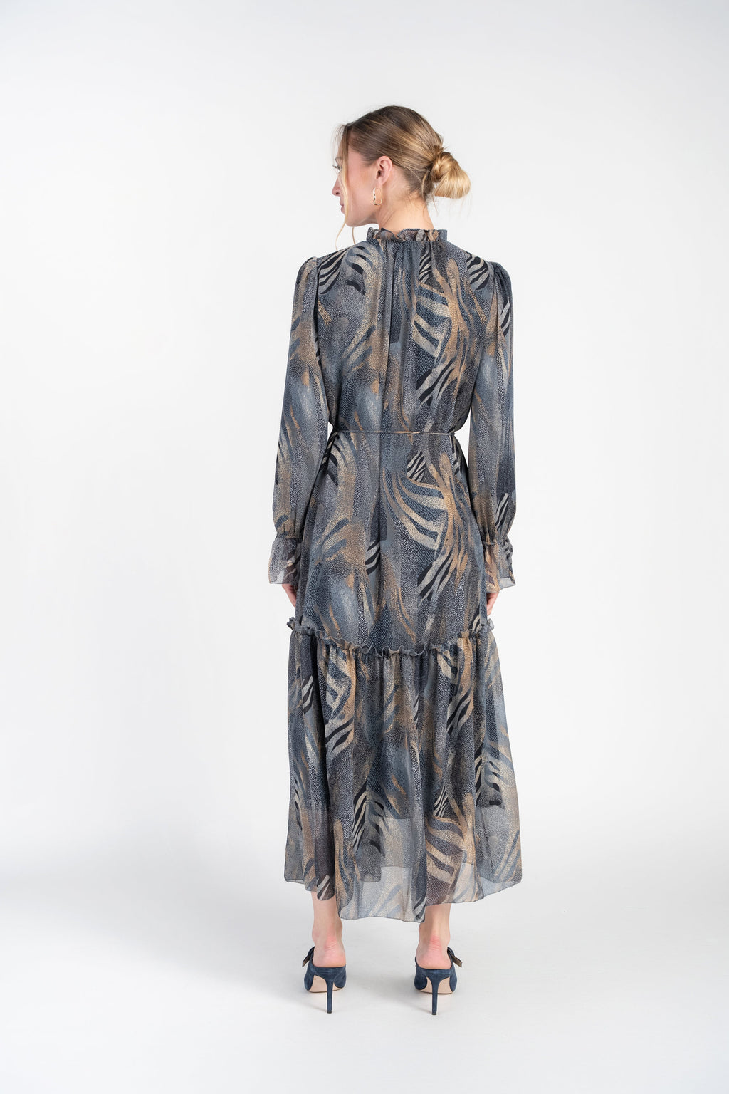 A woman viewed from the back wearing a long-sleeve midi dress with a gray and beige palm leaf pattern, paired with dark blue heels, against a plain white studio background.