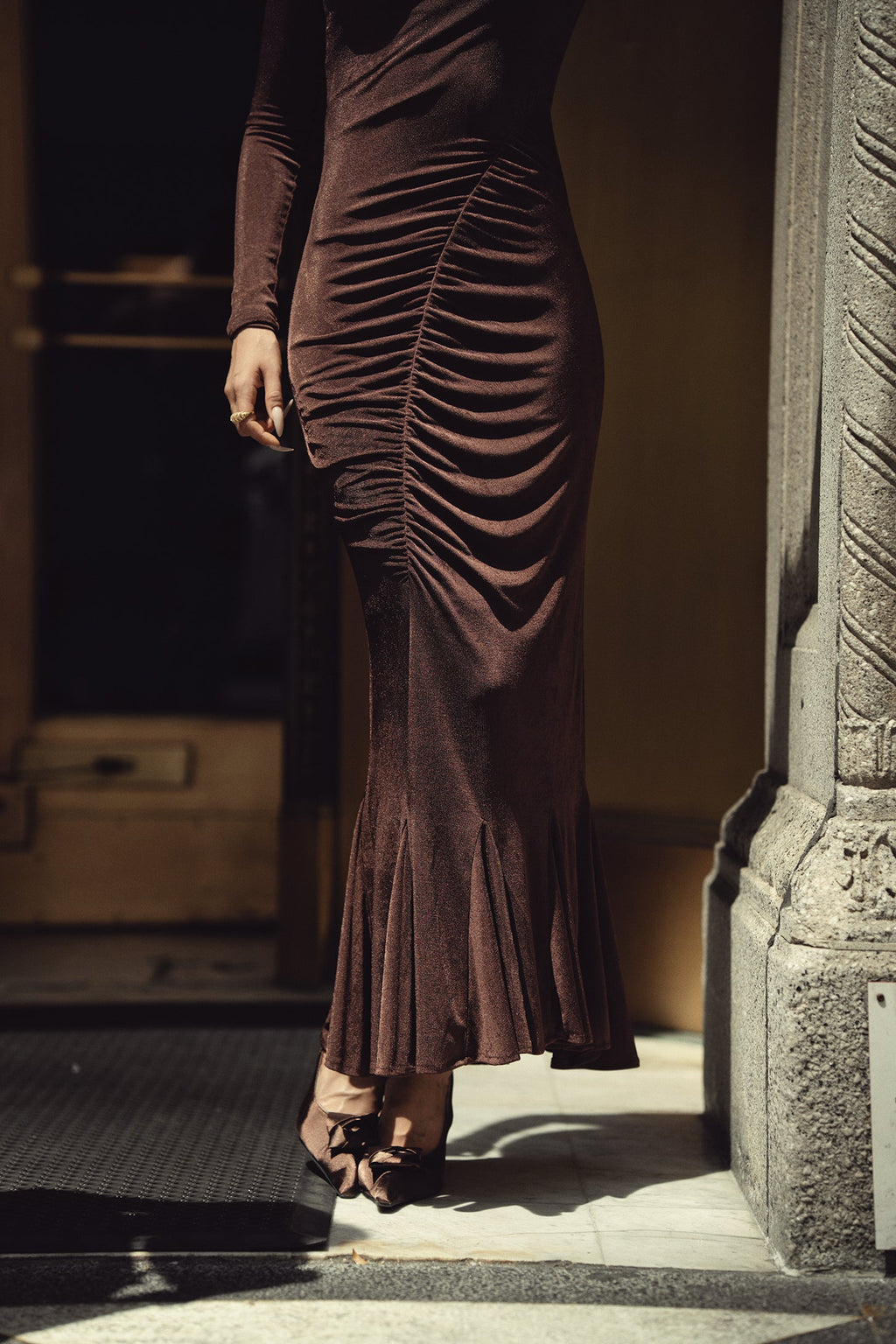A woman wearing a dark brown ruched long-sleeve maxi dress with a flared hem, paired with brown strappy heels, standing in a doorway.