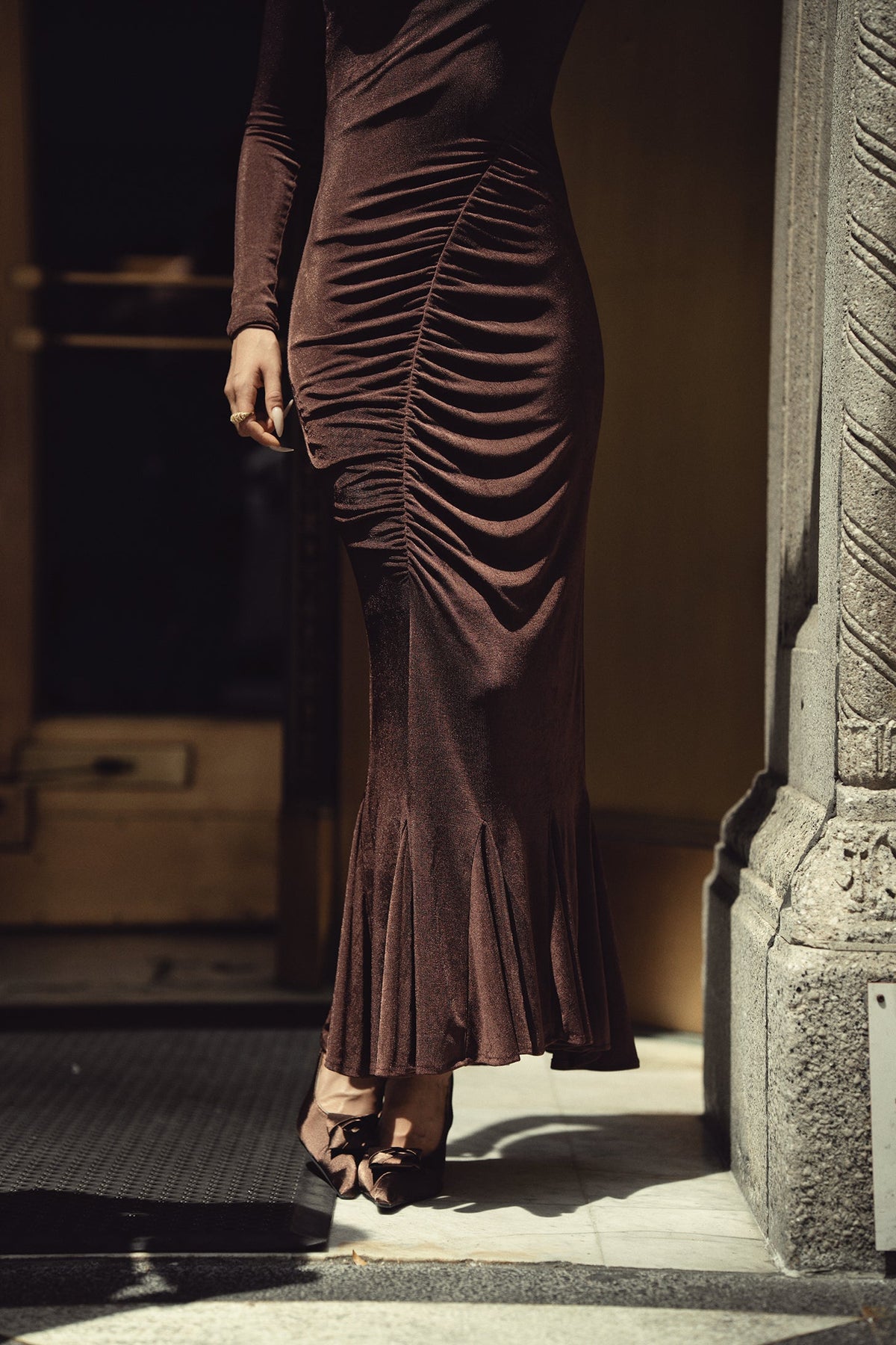 A woman wearing a dark brown ruched long-sleeve maxi dress with a flared hem, paired with brown strappy heels, standing in a doorway.