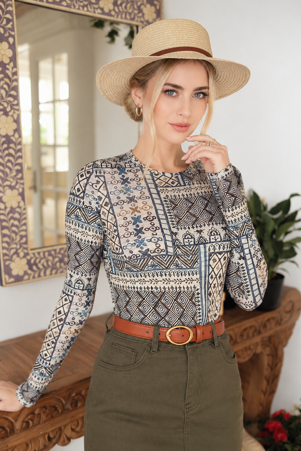 A woman wearing a long-sleeve crewneck top with a detailed geometric tribal pattern in beige, blue, and black, styled with an olive green skirt, a brown belt, and a straw hat.