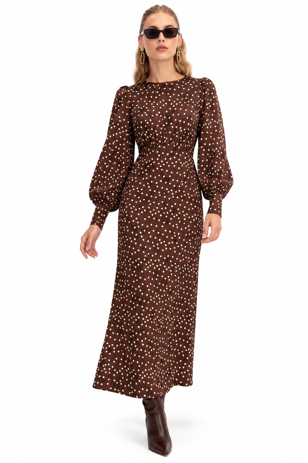 A woman modeling a long brown midi dress with a white polka dot pattern, featuring balloon sleeves and a gathered waist, paired with dark brown ankle boots and sunglasses against a plain white background.