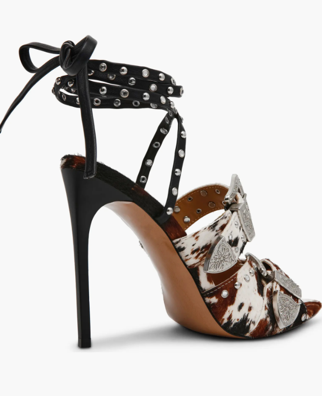 A single high-heeled sandal with a black studded ankle strap, a black stiletto heel, and a pointed toe. The footbed features a brown, black, and white cowhide print with large silver western-style conchos and studs.