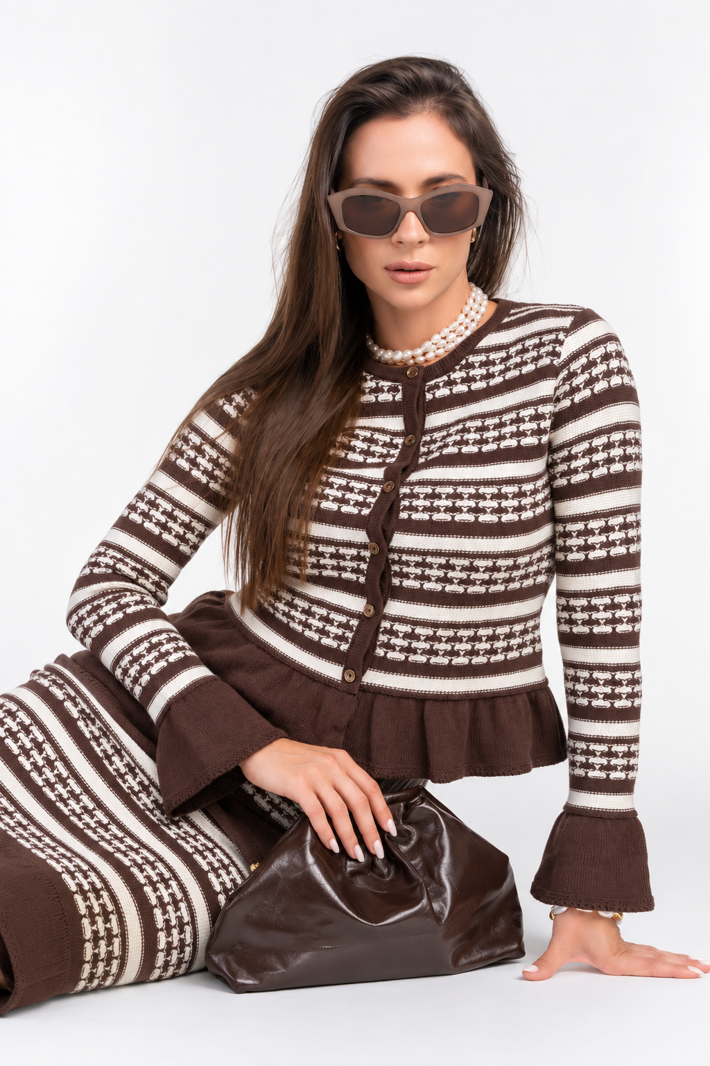 A woman models a matching brown and cream knit cardigan and skirt set with a geometric pattern, styled with sunglasses, a pearl necklace, and a brown handbag against a plain white background.