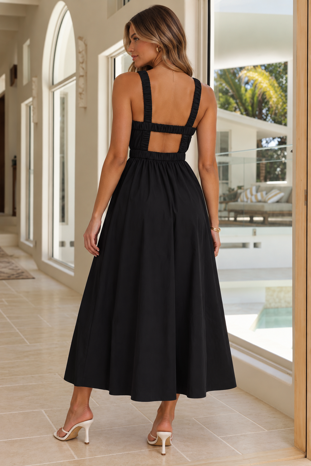 A woman wearing a black midi dress with a cutout back and wide shoulder straps, shown from behind in a bright indoor hallway with large windows.