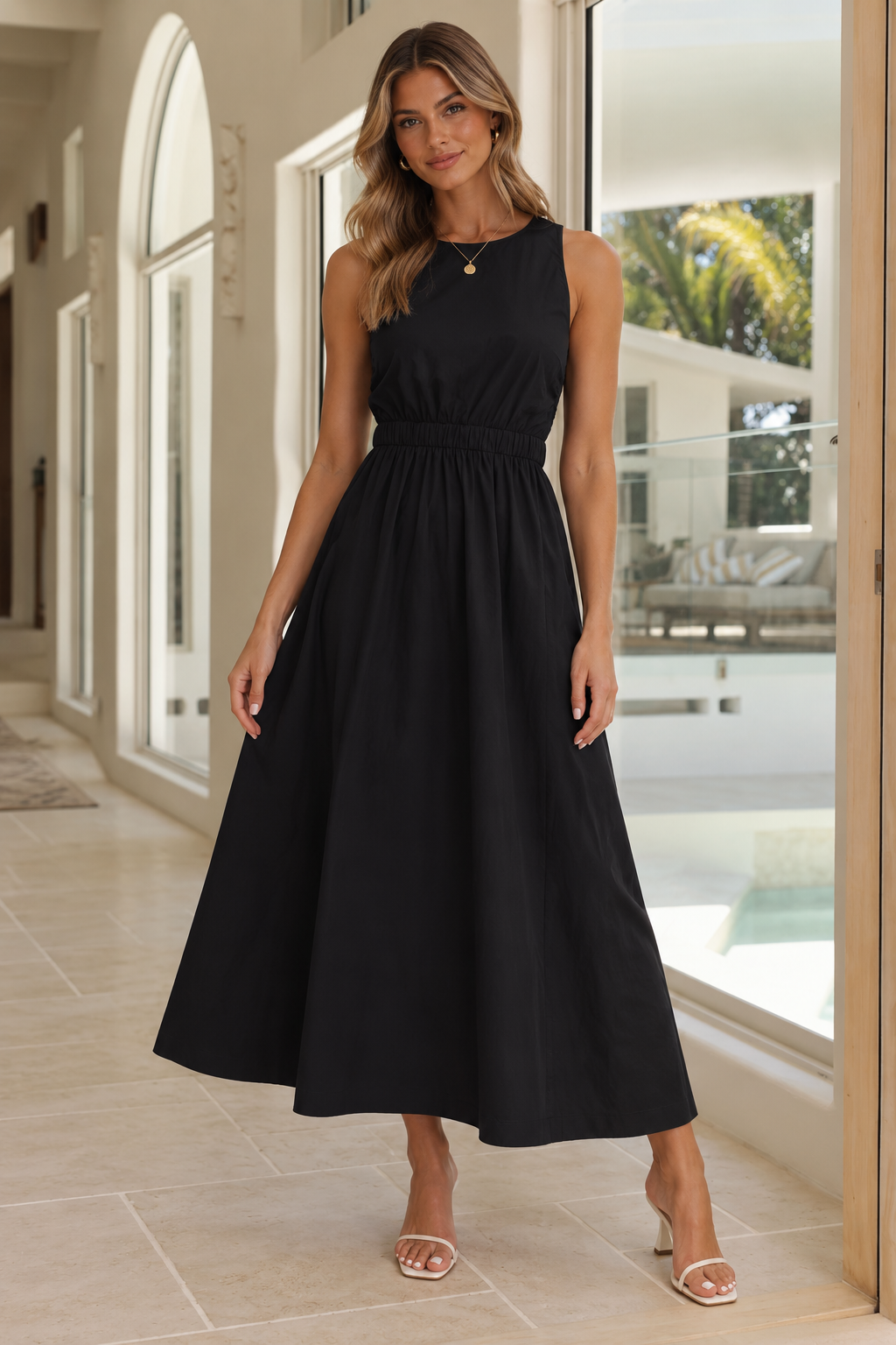 A woman modeling a sleeveless black midi dress with a gathered waist and flared skirt, standing in a bright, modern indoor hallway with large windows and tiled floors.
