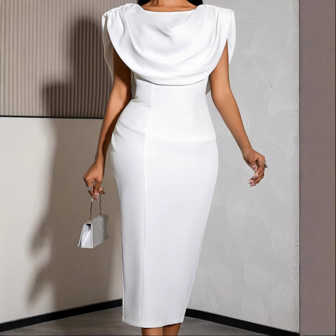 A woman wearing a white high-waisted midi dress with a draped cowl neckline and structured cap sleeves, holding a small metallic handbag in an indoor setting.