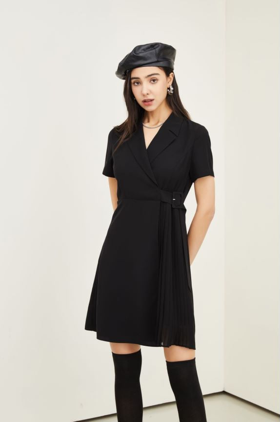 A woman modeling a black short-sleeve wrap-style dress with a tie waist, paired with a black beret and black knee-high socks, standing against a plain off-white wall.