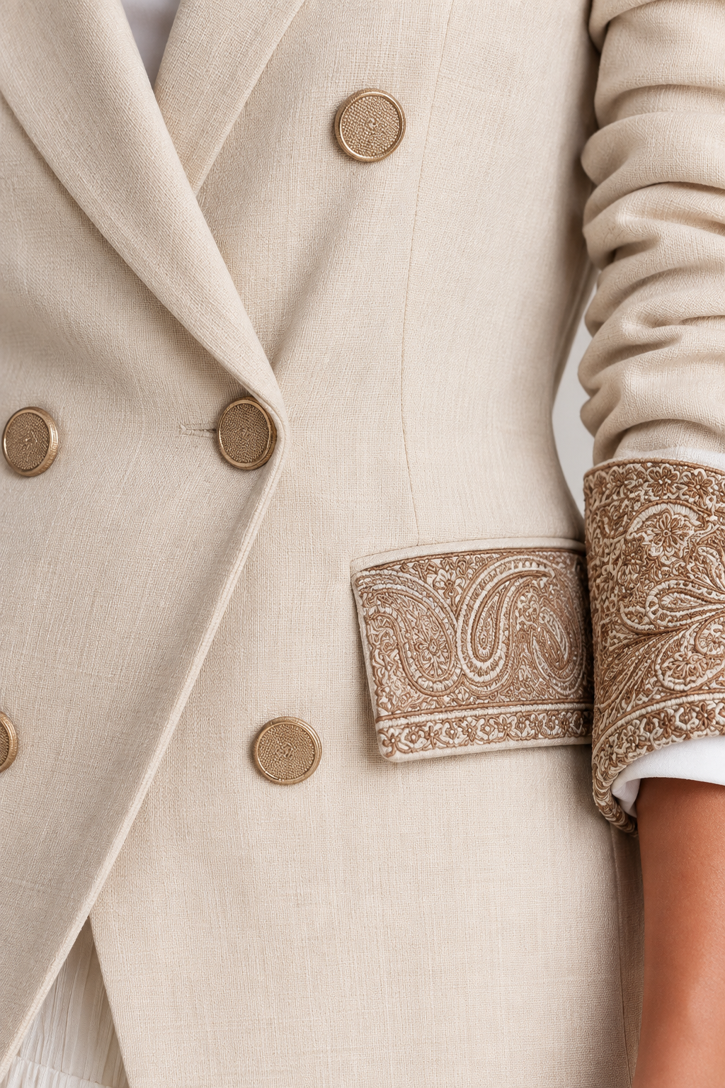 Close-up of a beige double-breasted blazer with gold buttons and ornate paisley embroidery on the pocket flap and cuffs, worn with a white undershirt.