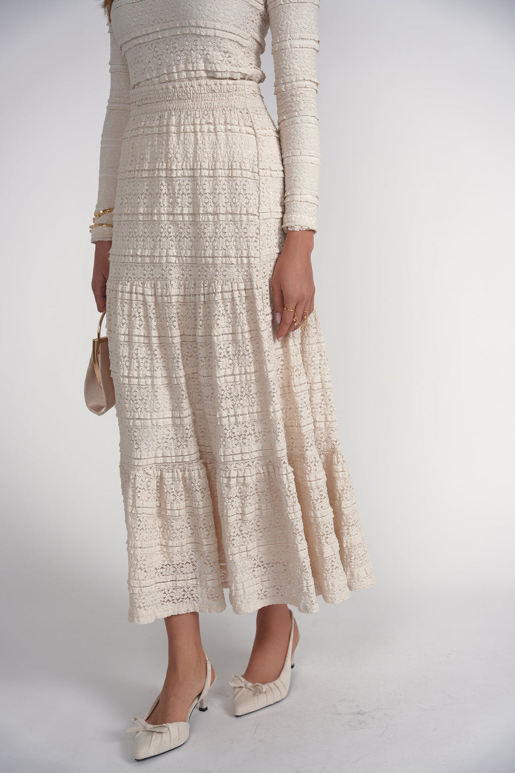 A model wearing a cream-colored textured lace maxi dress with long sleeves, paired with matching pointed-toe heels and holding a small beige handbag, set against a plain light gray studio background.