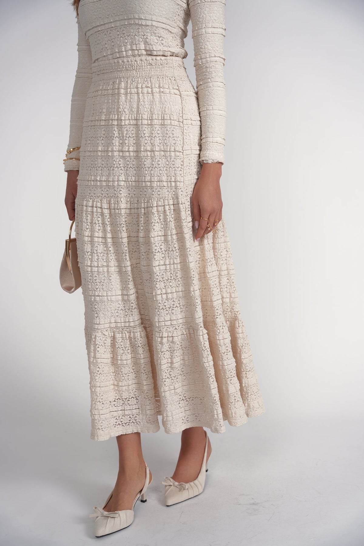 A model wearing a cream-colored textured lace maxi dress with long sleeves, paired with matching pointed-toe heels and holding a small beige handbag, set against a plain light gray studio background.