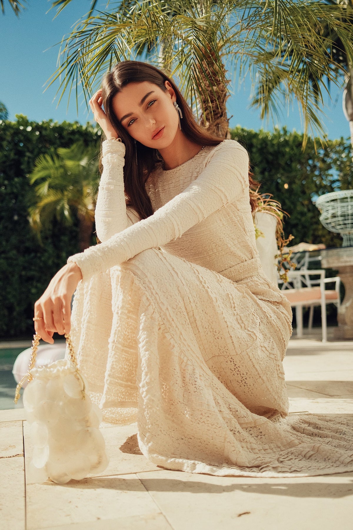 A woman wearing a long-sleeve cream lace dress sits outdoors on a tiled patio under palm trees, holding a white textured handbag.