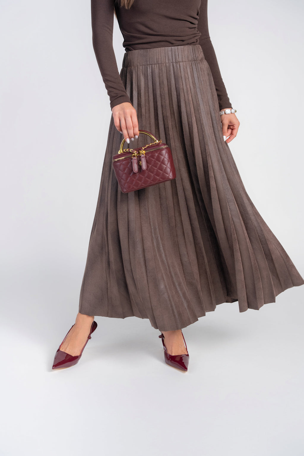 A model wearing a brown pleated maxi skirt, a matching brown long-sleeve top, burgundy pointed-toe heels, and holding a small burgundy quilted handbag against a light gray studio background.