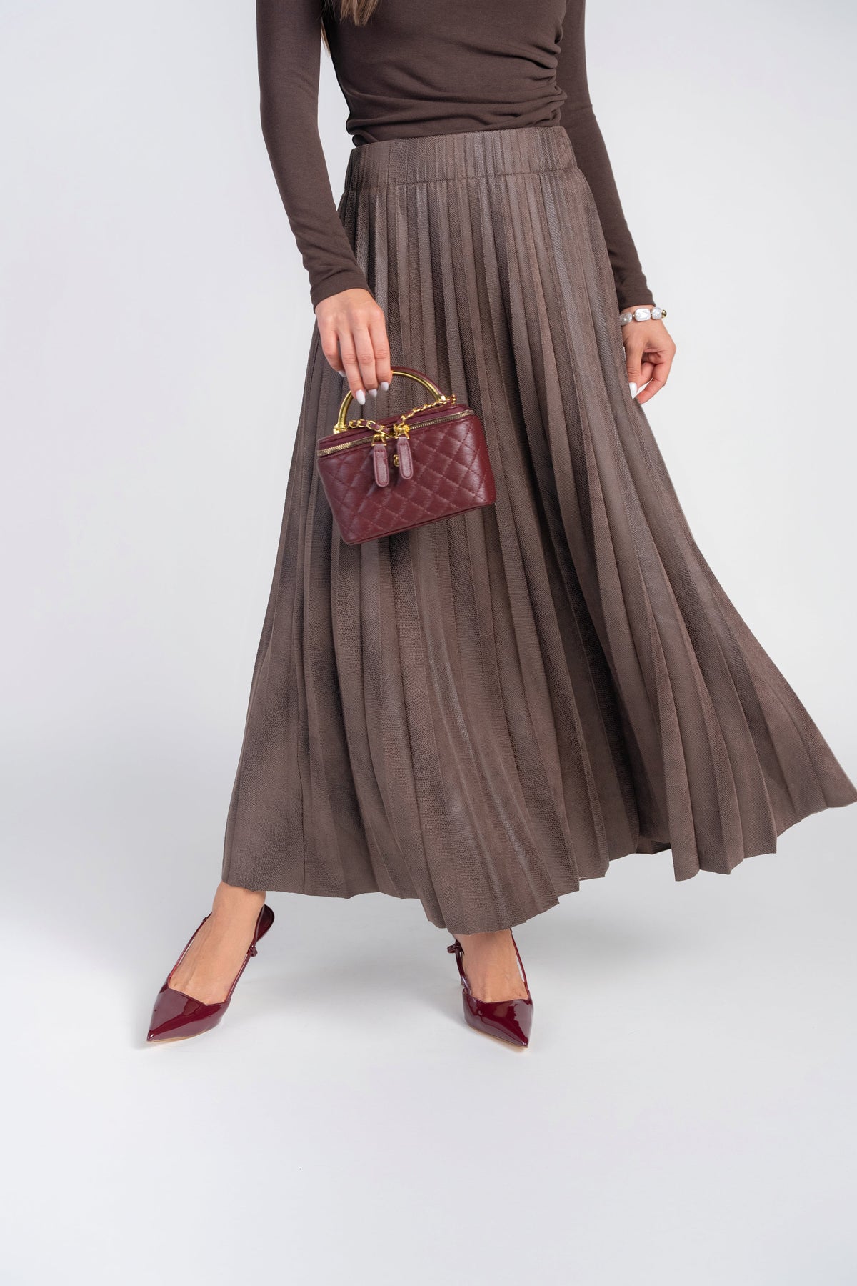 A model wearing a brown pleated maxi skirt, a matching brown long-sleeve top, burgundy pointed-toe heels, and holding a small burgundy quilted handbag against a light gray studio background.