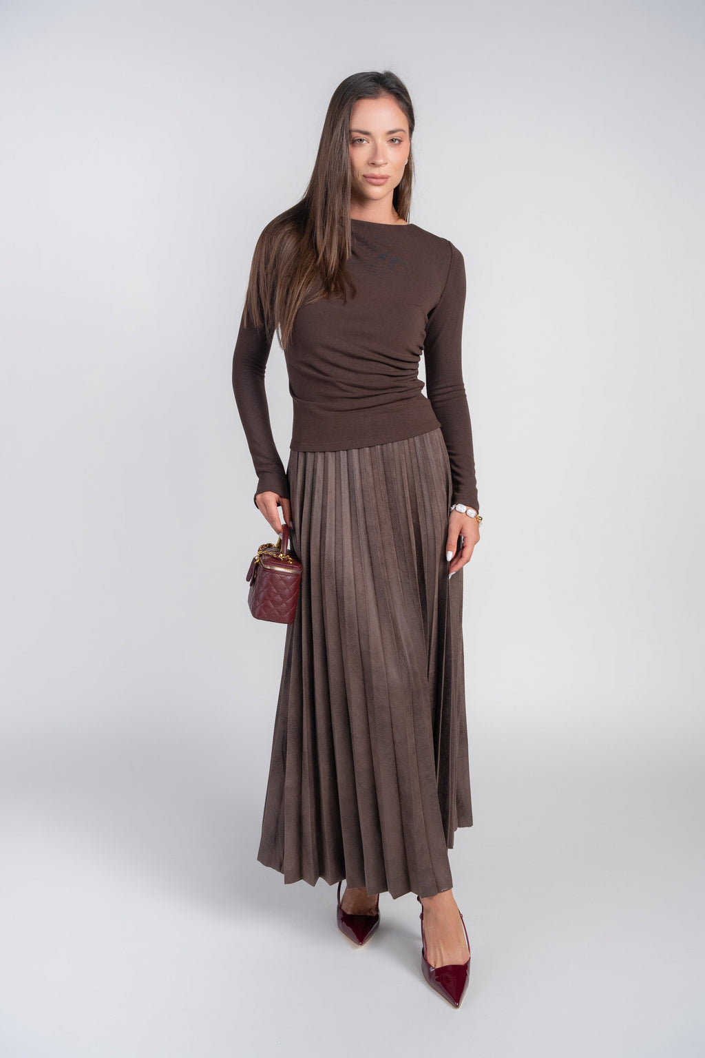 A woman modeling a dark brown long-sleeve top and matching pleated maxi skirt, holding a small dark red handbag and wearing dark red pointed-toe heels, against a light gray studio background.