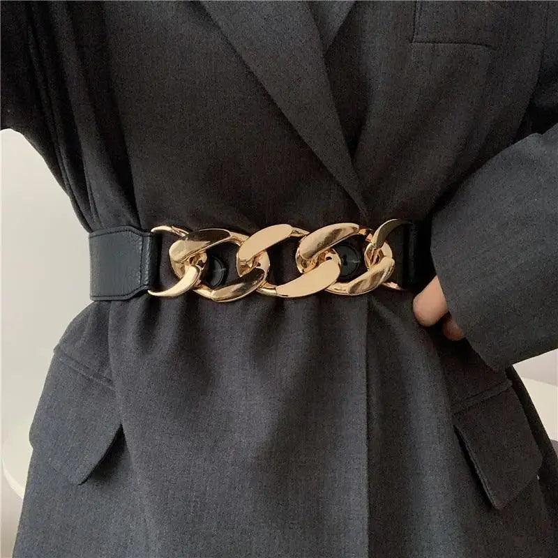 A close-up of a person wearing a wide black belt with oversized polished gold-tone chain links, styled over a dark gray blazer.
