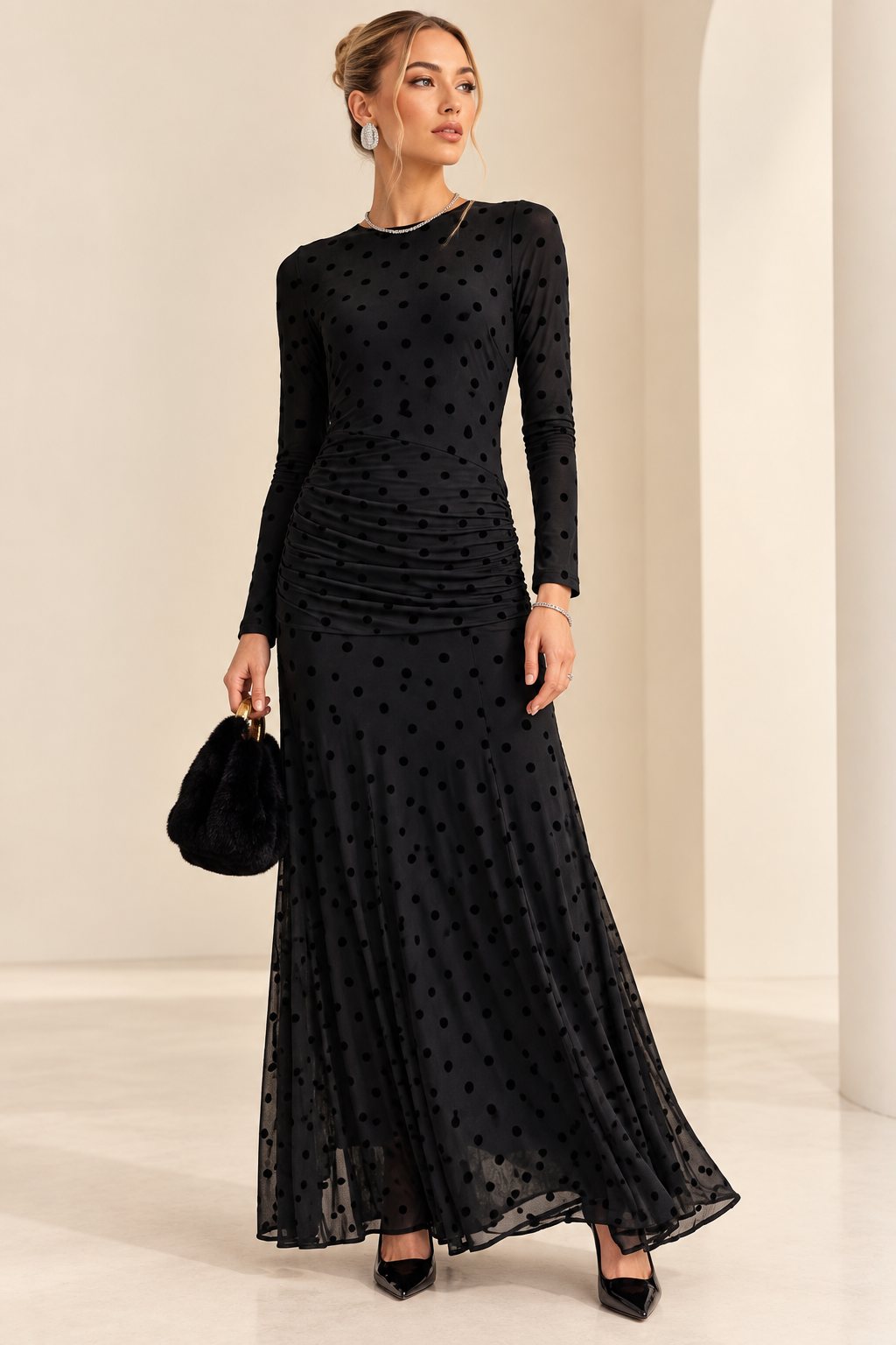 A woman models a long-sleeve black maxi dress with a polka dot pattern and ruched waist, holding a small black handbag in a minimalist cream-colored studio.