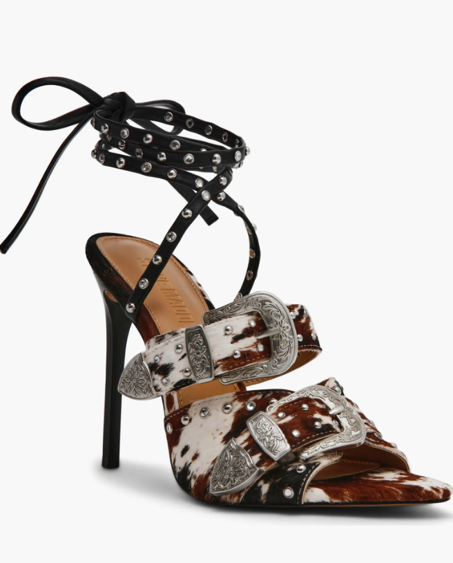 A single high-heeled stiletto sandal with a pointed toe, featuring brown and white cow-print straps with large silver concho-style buckles, a black studded ankle strap, and a black heel and toe post, shown on a light gray background.