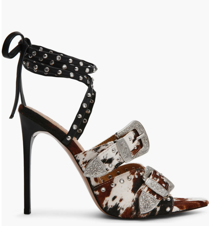 A high-heeled stiletto sandal with a brown and white cow-print upper, black ankle straps, and silver-tone western-style buckles and studs, shown against a plain white background.