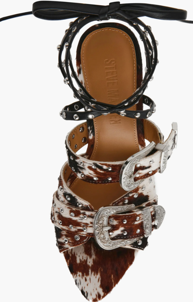 A single pointed-toe heeled sandal with a brown and white cowhide pattern, adorned with silver studs, western-style buckles, and black lace-up ankle straps, shown against a plain white background.