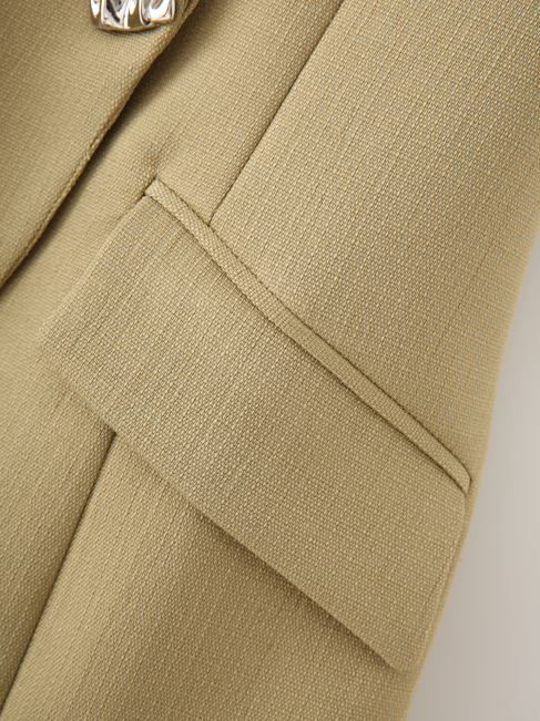 Close-up of a beige textured fabric garment, showing a folded pocket flap and part of a metallic button.